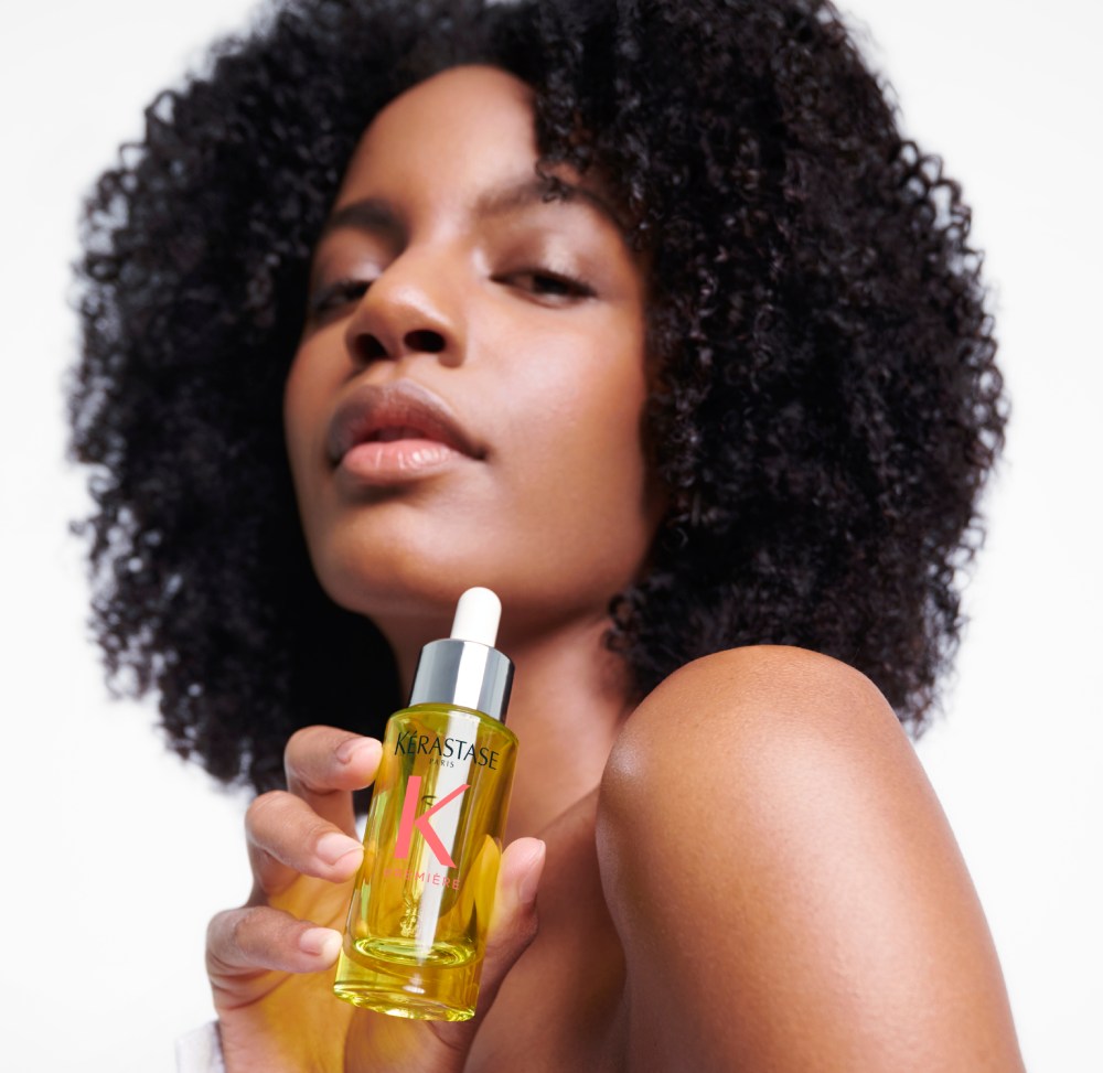 Ebonee Davis, com seu lindo cabelo cacheado, segura o melhor óleo capilar para cabelos danificados Première Huile Gloss Réparatrice.